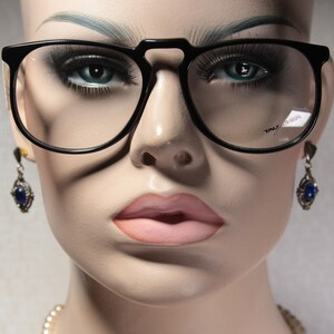 May include: Black-framed eyeglasses with a distinctive bridge design. The glasses are on a mannequin head, paired with blue-stoned earrings. The mannequin has full makeup, including blue eyeshadow and pink lipstick. The glasses have a brand name on the lens.
