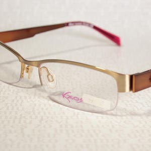 Peut inclure: Lunettes semi-cerclées avec une monture en métal doré et des branches marron. Le verre droit porte l'inscription rose Kaos. Les branches présentent une finition marron avec un détail découpé, et l'intérieur des embouts est rose vif.