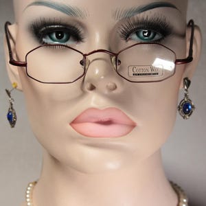 Könnte beinhalten: Eine Schaufensterpuppe mit einer bordeauxfarbenen Brille mit rechteckiger Form und dem Text "COTTON WAY" auf dem Glas. Die Schaufensterpuppe hat auch blaue Edelsteinohrringe und eine Perlenkette.