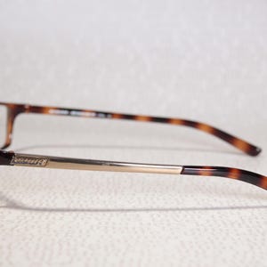 May include: Rectangular eyeglasses with a brown tortoiseshell pattern on the frames and temples. The arms have a gold-coloured metal section. The lenses are clear. The glasses are set against a neutral background.