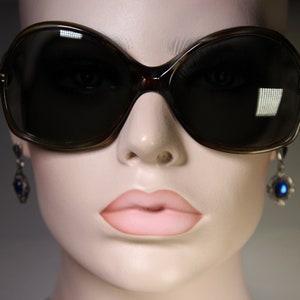 K&ouml;nnte beinhalten: &Uuml;bergro&szlig;e, dunkelbraune Sonnenbrille mit Schildpattmuster. Die Sonnenbrille hat eine gro&szlig;e, abgerundete Form und dunkle Gl&auml;ser. Das Bild zeigt auch eine Schaufensterpuppe, die Ohrringe mit blauen Edelsteinen tr&auml;gt.