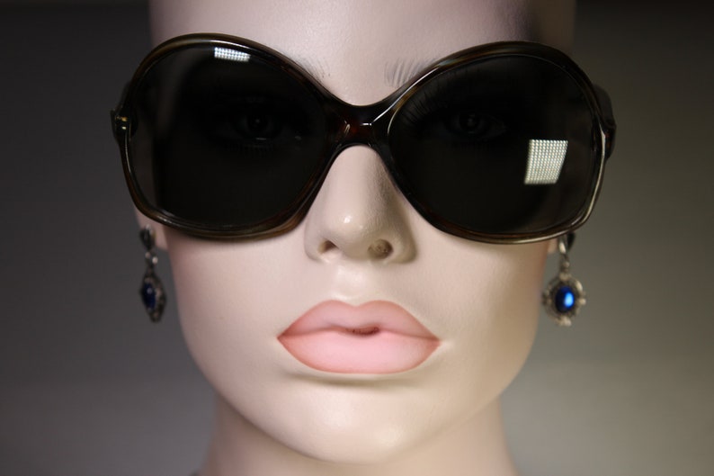 K&ouml;nnte beinhalten: &Uuml;bergro&szlig;e, dunkelbraune Sonnenbrille mit Schildpattmuster. Die Sonnenbrille hat eine gro&szlig;e, abgerundete Form und dunkle Gl&auml;ser. Das Bild zeigt auch eine Schaufensterpuppe, die Ohrringe mit blauen Edelsteinen tr&auml;gt.