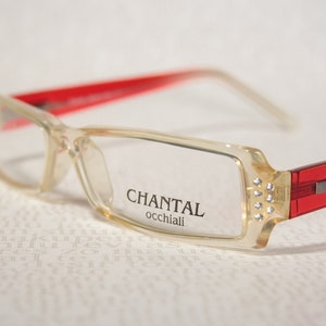 Può includere: Occhiali con montatura in plastica trasparente, aste rosse e strass sul lato. Gli occhiali hanno la scritta "CHANTAL occhiali" stampata sulla montatura.