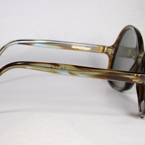 K&ouml;nnte beinhalten: Vintage Sonnenbrille mit einem braunen und beigefarbenen marmorierten Rahmen und dunkelgrauen Gl&auml;sern. Die B&uuml;gel der Sonnenbrille haben ein hellblaues und braunes Muster. Der Markenname ist auf dem B&uuml;gel aufgedruckt. Ein klassisches Accessoire.