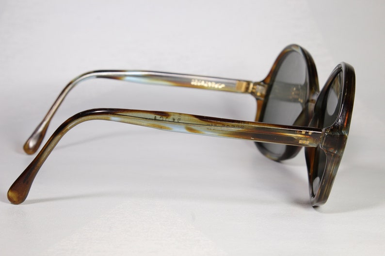 K&ouml;nnte beinhalten: Vintage Sonnenbrille mit einem braunen und beigefarbenen marmorierten Rahmen und dunkelgrauen Gl&auml;sern. Die B&uuml;gel der Sonnenbrille haben ein hellblaues und braunes Muster. Der Markenname ist auf dem B&uuml;gel aufgedruckt. Ein klassisches Accessoire.