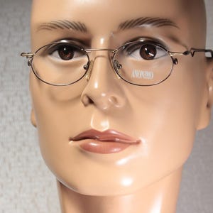 Op de afbeelding: Ovale bril met een dun, goudkleurig metalen montuur. De glazen zijn helder en de tekst "ANONIMO" is zichtbaar op het rechterglas. De bril wordt getoond op een mannequinhoofd.