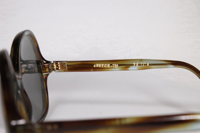 K&ouml;nnte beinhalten: Vintage Sonnenbrille mit Schildpattmuster und dunklen Gl&auml;sern. Der Rahmen hat ein klassisches Design mit einem Farbverlauf aus Braun und Beige. Der Text "GREECE-TM" und "52 [] 14" ist auf dem B&uuml;gel aufgedruckt.