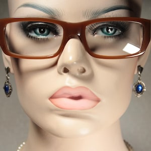 Op de afbeelding: Een mannequinhoofd met bruine rechthoekige bril en blauwe edelsteen oorbellen. De mannequin heeft een lichte huid, volle lippen en lange wimpers. De achtergrond is een neutraal grijs.