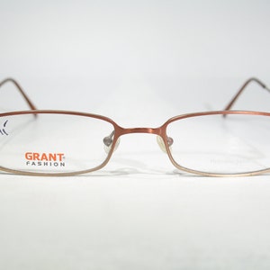 Op de afbeelding: Een paar rechthoekige metalen brillen in bruin met heldere glazen. De merknaam "GRANT FASHION" staat op de linkerlens gedrukt.