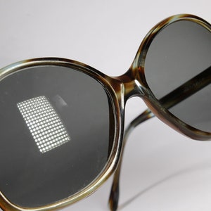 K&ouml;nnte beinhalten: &Uuml;bergro&szlig;e Sonnenbrille mit einem Rahmen im Schildpattmuster und dunkelgrauen Gl&auml;sern. Die Brille hat ein klassisches Retro-Design mit einem dicken Rahmen und einer leichten Cat-Eye-Form. Die Gl&auml;ser reflektieren ein Lichtraster.