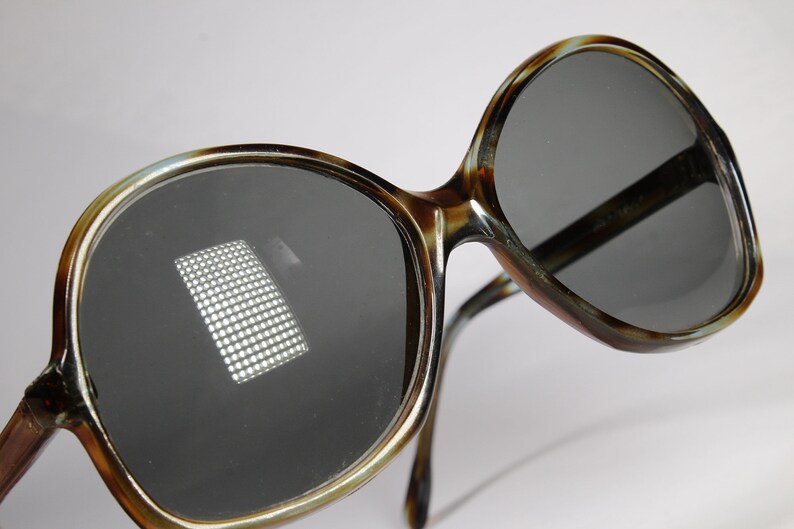 K&ouml;nnte beinhalten: &Uuml;bergro&szlig;e Sonnenbrille mit einem Rahmen im Schildpattmuster und dunkelgrauen Gl&auml;sern. Die Brille hat ein klassisches Retro-Design mit einem dicken Rahmen und einer leichten Cat-Eye-Form. Die Gl&auml;ser reflektieren ein Lichtraster.