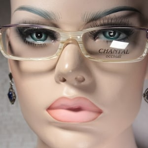 Pode incluir: Um close-up de um manequim usando óculos bege e roxo com o nome da marca "CHANTAL occhiali" impresso na lente direita. O manequim também está usando brincos de pedras preciosas azuis.