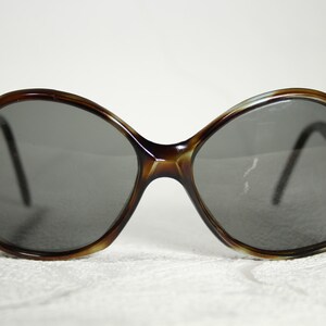 May include: A pair of brown tortoiseshell sunglasses with large, round lenses. The lenses are dark grey and the frames are made of plastic.
