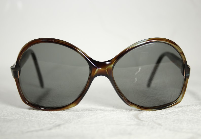 May include: A pair of brown tortoiseshell sunglasses with large, round lenses. The lenses are dark grey and the frames are made of plastic.