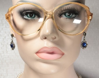 May include: A pair of large, round, clear, plastic eyeglasses with gold frames. The eyeglasses are on a mannequin's face. The mannequin is wearing blue gemstone earrings.