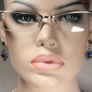 May include: A silver-toned pair of eyeglasses with clear lenses. The eyeglasses have a thin, rectangular frame with a slight curve at the top. The model is wearing blue gemstone earrings.
