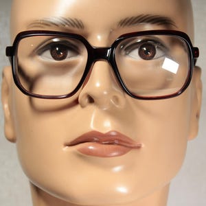 Op de afbeelding: Close-up van een mannequinhoofd met een grote, vierkant omrande bril. De monturen zijn donker bordeauxrood van kleur en de glazen zijn helder. De mannequin heeft bruine ogen en een neutrale huidskleur.