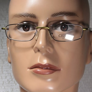 May include: A mannequin wearing gold-rimmed glasses with clear lenses. The glasses have the text "CHARMANT" on the right lens.