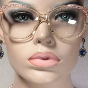Könnte beinhalten: Nahaufnahme eines Mannequins, das eine große, klare, plastikgefasste Brille mit einem braunen und weißen Muster trägt. Das Mannequin trägt auch ein Paar baumelnde Ohrringe mit blauen Edelsteinen.