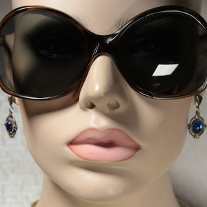 May include: A close-up of a mannequin wearing large, brown, oval-shaped sunglasses with dark lenses. The mannequin is also wearing silver dangle earrings with blue gemstones.