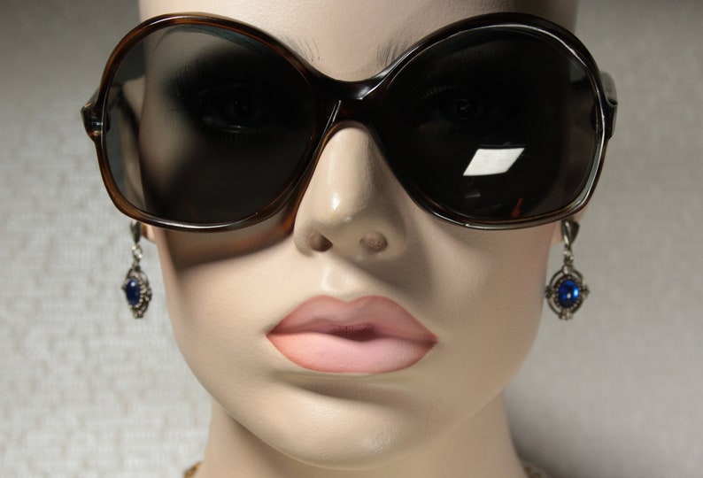 May include: A close-up of a mannequin wearing large, brown, oval-shaped sunglasses with dark lenses. The mannequin is also wearing silver dangle earrings with blue gemstones.