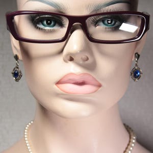 Op de afbeelding: Een close-up van een vrouwengezicht met een donkerpaarse bril met een zilveren montuur en zilveren oorbellen met blauwe edelstenen.