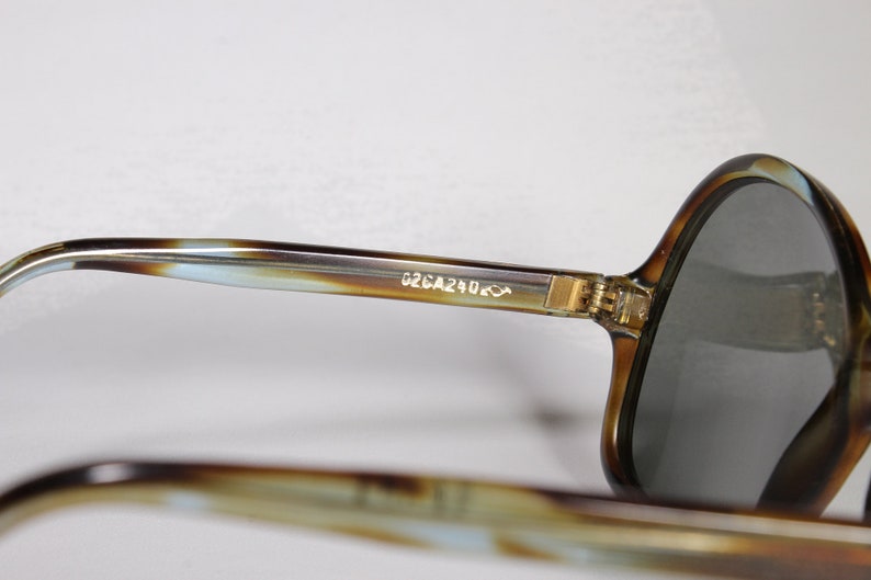K&ouml;nnte beinhalten: Nahaufnahme einer Vintage-Sonnenbrille mit Schildpattmuster. Die Fassungen sind braun und beige mit einem hellblauen Akzent. Die Gl&auml;ser sind grau. Der Text "G2CA240" ist auf dem B&uuml;gel aufgedruckt.