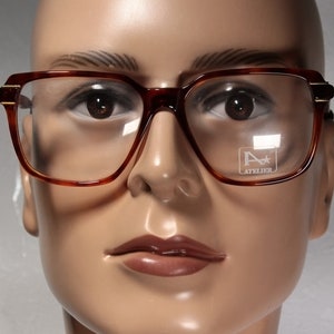 Könnte beinhalten: Braune Brille mit quadratischem Rahmen und goldenen Akzenten an den Bügeln. Die Brille hat klare Gläser und der Text "A ATELIER" ist auf einem der Gläser zu sehen. Die Fassungen sind in einem warmen, rötlich-braunen Farbton gehalten.