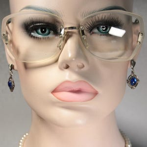 May include: Oversized eyeglasses with translucent beige frames and clear lenses, featuring a gold-tone metal bridge and hinges. The glasses are paired with dangling earrings that have a silver-tone setting and a central oval blue stone.