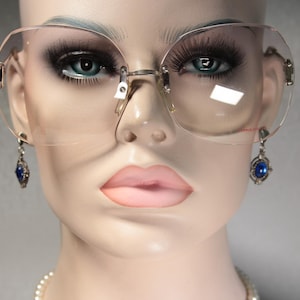 May include: Rimless spectacles with large, gradient-tinted lenses and thin gold-tone metal temples. The glasses feature a bridge with adjustable nose pads and small, dangling silver-tone earrings with blue stones attached to the frame hinges.