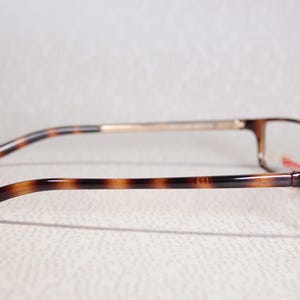 May include: Rectangular eyeglasses with a tortoiseshell pattern. The frames are brown and black. The temples have a similar pattern and connect to a gold-coloured bridge. The lenses are clear.
