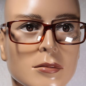 May include: Brown rectangular eyeglasses with clear lenses. The glasses have a small logo on the right lens that says "OPTICA".
