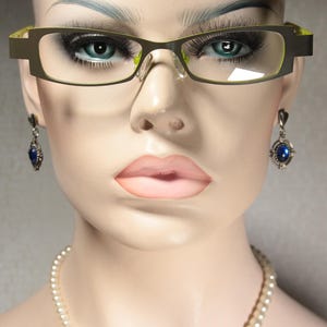 Pode incluir: Uma mulher usando óculos de armação verde com brincos de pedras preciosas azuis. Ela está usando um colar de pérolas.