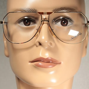 May include: Gold-rimmed aviator eyeglasses with clear lenses. The glasses have a double bridge and a rectangular shape. The lenses have the text "John Lennon" printed on them. The glasses are displayed on a mannequin.