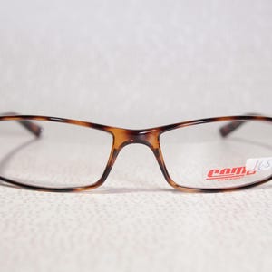 May include: Tortoiseshell pattern eyeglasses with clear rectangular lenses. The brand name "com" is printed in red on the lower right lens. The frame has a classic design. The glasses are suitable for everyday use.