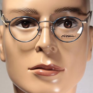May include: Round eyeglasses with a thin silver metal frame and blue patterned rims. The right lens features the text titan and kevytykset printed in black. The glasses are positioned on a beige mannequin head with brown eyes and neutral features.