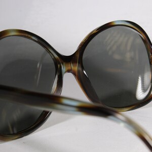 K&ouml;nnte beinhalten: &Uuml;bergro&szlig;e Sonnenbrille mit einem Rahmen im Schildpattmuster und dunkelgrauen Gl&auml;sern. Die Sonnenbrille hat einen Retro-Vintage-Stil mit einer breiten, abgerundeten Form. Die B&uuml;gel haben eine &auml;hnliche Farbe wie der Rahmen. Die Gl&auml;ser sind reflektierend.