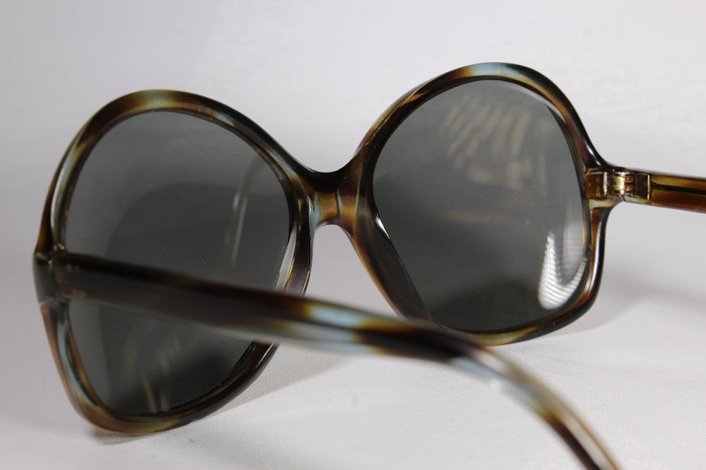 K&ouml;nnte beinhalten: &Uuml;bergro&szlig;e Sonnenbrille mit einem Rahmen im Schildpattmuster und dunkelgrauen Gl&auml;sern. Die Sonnenbrille hat einen Retro-Vintage-Stil mit einer breiten, abgerundeten Form. Die B&uuml;gel haben eine &auml;hnliche Farbe wie der Rahmen. Die Gl&auml;ser sind reflektierend.