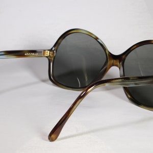 K&ouml;nnte beinhalten: Sonnenbrille mit Schildpattmuster und dunkelgrauen Gl&auml;sern. Die Fassungen haben ein braunes, beiges und blaues Muster. Die B&uuml;gel der Sonnenbrille sind gebogen und haben ein &auml;hnliches Muster. Die Sonnenbrille ist abgewinkelt.