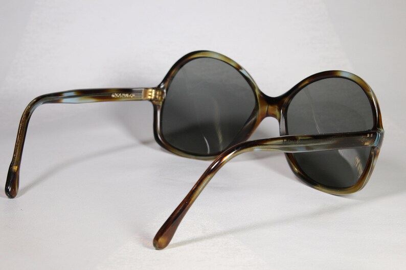 K&ouml;nnte beinhalten: Sonnenbrille mit Schildpattmuster und dunkelgrauen Gl&auml;sern. Die Fassungen haben ein braunes, beiges und blaues Muster. Die B&uuml;gel der Sonnenbrille sind gebogen und haben ein &auml;hnliches Muster. Die Sonnenbrille ist abgewinkelt.