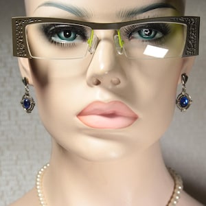 Peut inclure: Un mannequin portant des lunettes à monture argentée avec un design décoratif, des boucles d'oreilles en pierres précieuses bleues et un collier de perles.