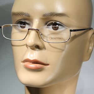 Könnte beinhalten: Nahaufnahme eines Mannequins, das eine dünne, rechteckige Brille mit silbernem Rahmen und klaren Gläsern trägt. Die Brille hat ein "Nickel Free"-Etikett auf der rechten Linse.
