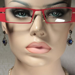 Puede incluir: Un maniquí con gafas de montura roja con lentes transparentes y pendientes de piedras preciosas azules.