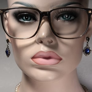 May include: A close-up of a mannequin wearing brown-framed glasses with clear lenses and blue gemstone earrings. The mannequin is wearing a pearl necklace.