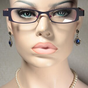 May include: A mannequin wearing black framed glasses with pink accents, a pearl necklace, and blue gemstone earrings.