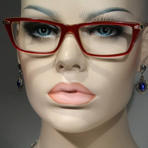 Könnte beinhalten: Eine Mannequin mit einer roten Brille mit goldenen Akzenten und blauen Edelsteinohrringen.