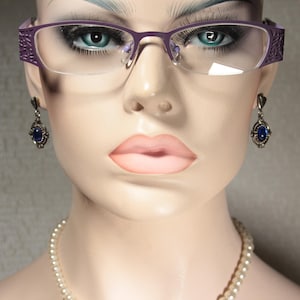 Peut inclure: Un mannequin portant des lunettes violettes avec un collier de perles et des boucles d'oreilles en pierres précieuses bleues.