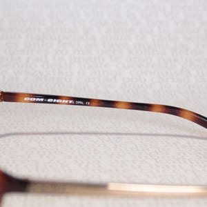 May include: Close-up of eyeglasses with a brown tortoiseshell pattern on the arms. The text "COM-EIGHT, OPAL CE" is printed on the arm. The glasses have a rectangular frame.