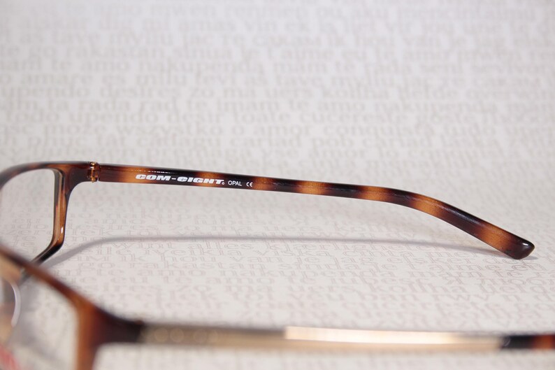 May include: Close-up of eyeglasses with a brown tortoiseshell pattern on the arms. The text "COM-EIGHT, OPAL CE" is printed on the arm. The glasses have a rectangular frame.