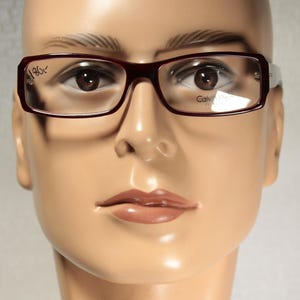 May include: Rectangular eyeglasses with a burgundy frame and clear lenses. The glasses have the brand name "Calvin Klein" printed on the lens. The frame has a white accent on the temples. The glasses are displayed on a mannequin head.